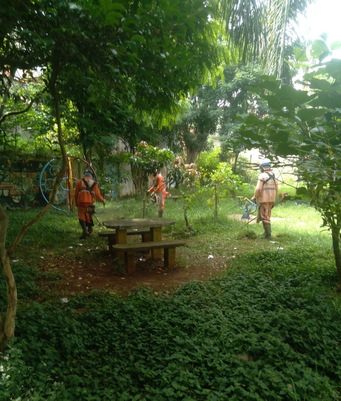 A imagem mostra três trabalhadores da prefeitura com roupas laranja usando equipamentos de proteção enquanto fazem a manutenção de um jardim. Eles estão cortando a grama e cuidando das plantas ao redor de uma mesa de concreto com bancos, em uma área verde cercada por árvores.